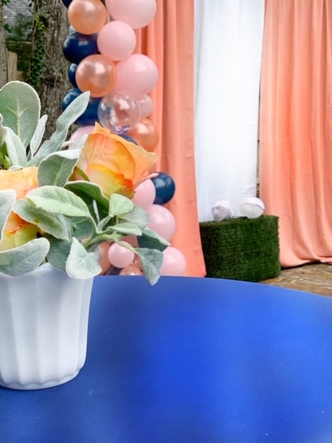 blue covered table in forground with white milk glass vase that is holding coral roses and silver lambs ear. in the background is two coral drapes with a white drape in the middle photo our photo spot.